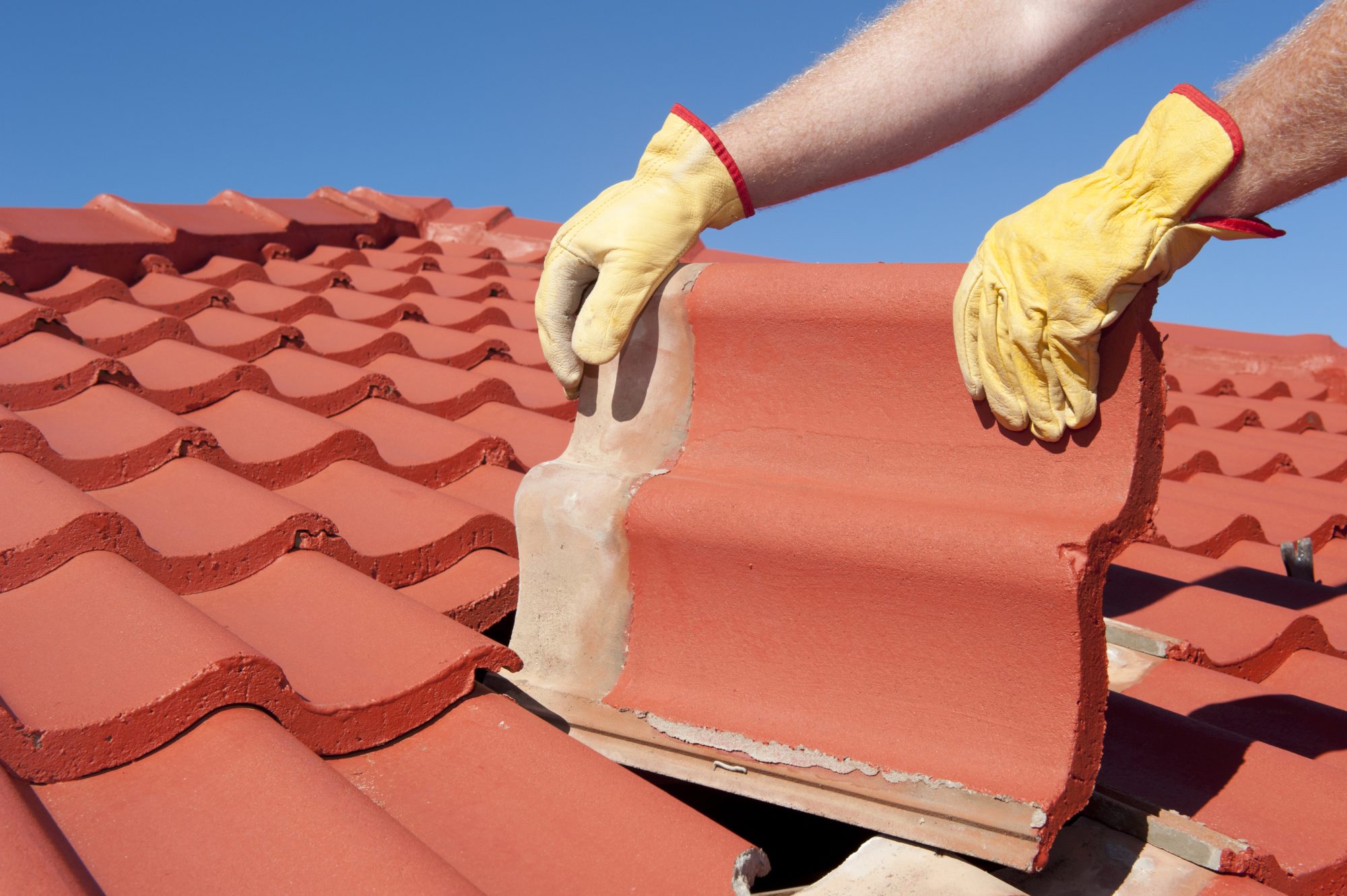 Roof repairs, worker with yellow gloves replacing red tiles or shingles on house with blue sky as background and copy space.